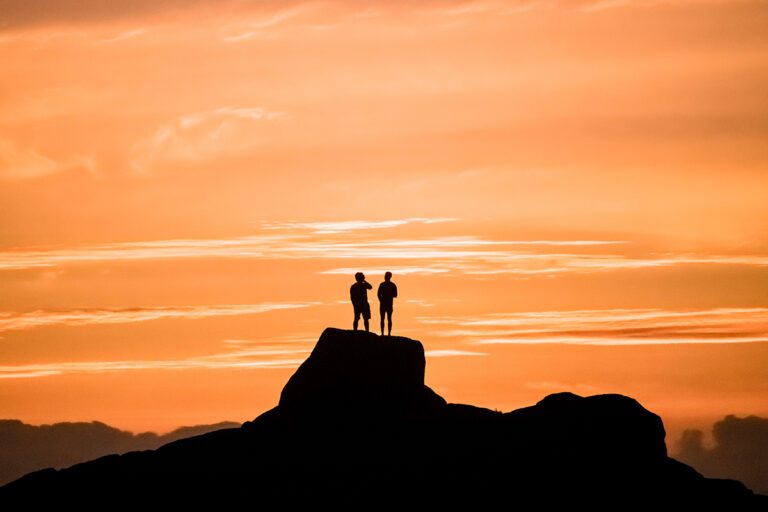 Dramatic golden sunset of two men admiring the view on vacation, standing on a large rock on a beach in Jacobsbaai, South Africa.